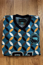 Load image into Gallery viewer, A folded V-neck knit sweater from Washington Motorsports on a wooden floor. The sweater features a teal blue background with a repeating geometric pattern of black, white, brown, and orange ovals.
